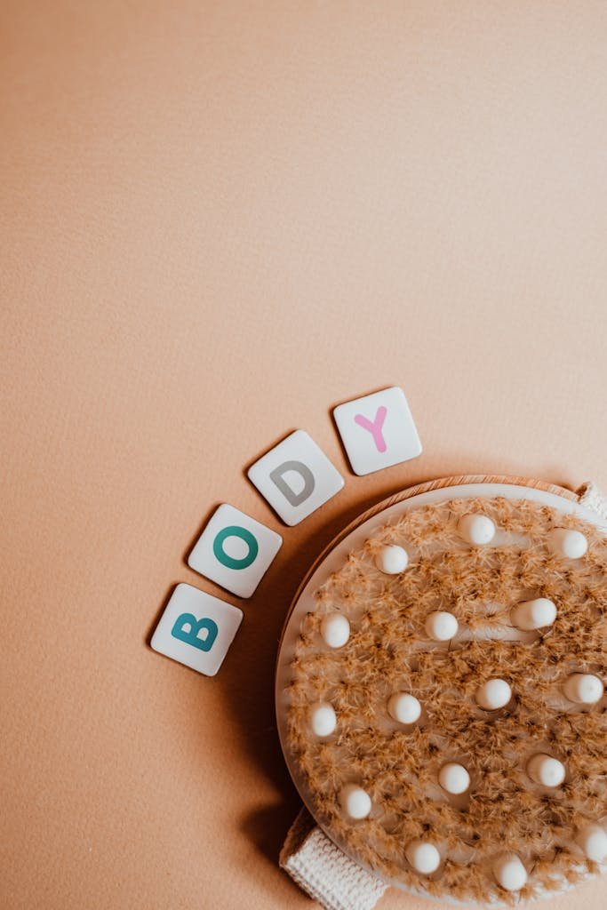 A flat lay of an exfoliating dry body brush paired with letter cubes spelling 'BODY' on a beige background.