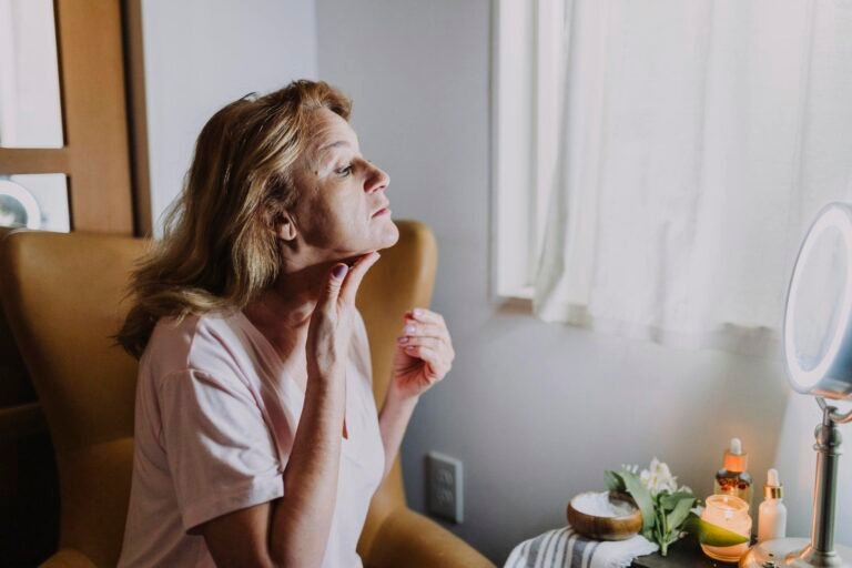 A mature woman examining her skin in a well-lit room with skincare products nearby.