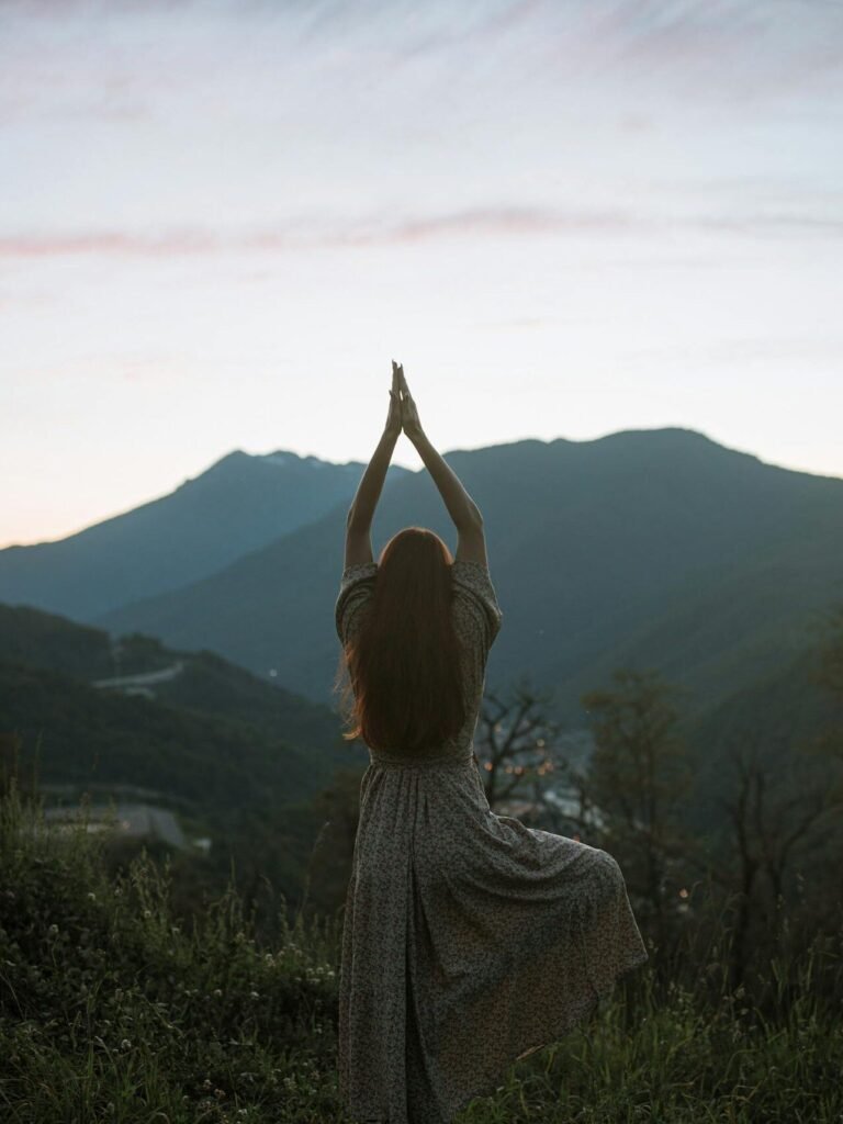 A woman in a dress performs tree pose against a mountainous sunrise backdrop, adding serenity and strength.