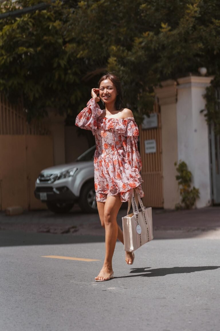 Barefoot woman in a floral dress walking on a sunny street, exuding carefree vibes with a tote bag.