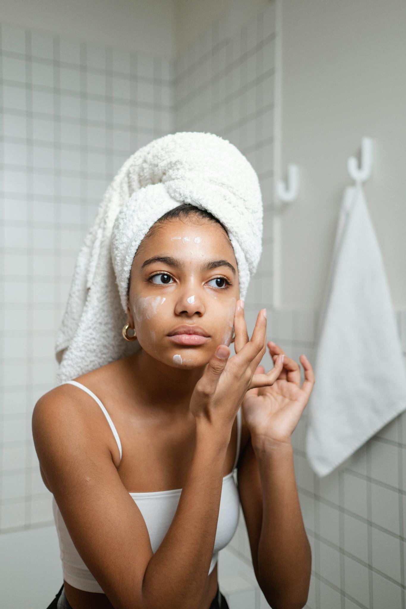Young woman with a towel on her head applying skincare cream in a modern bathroom.
