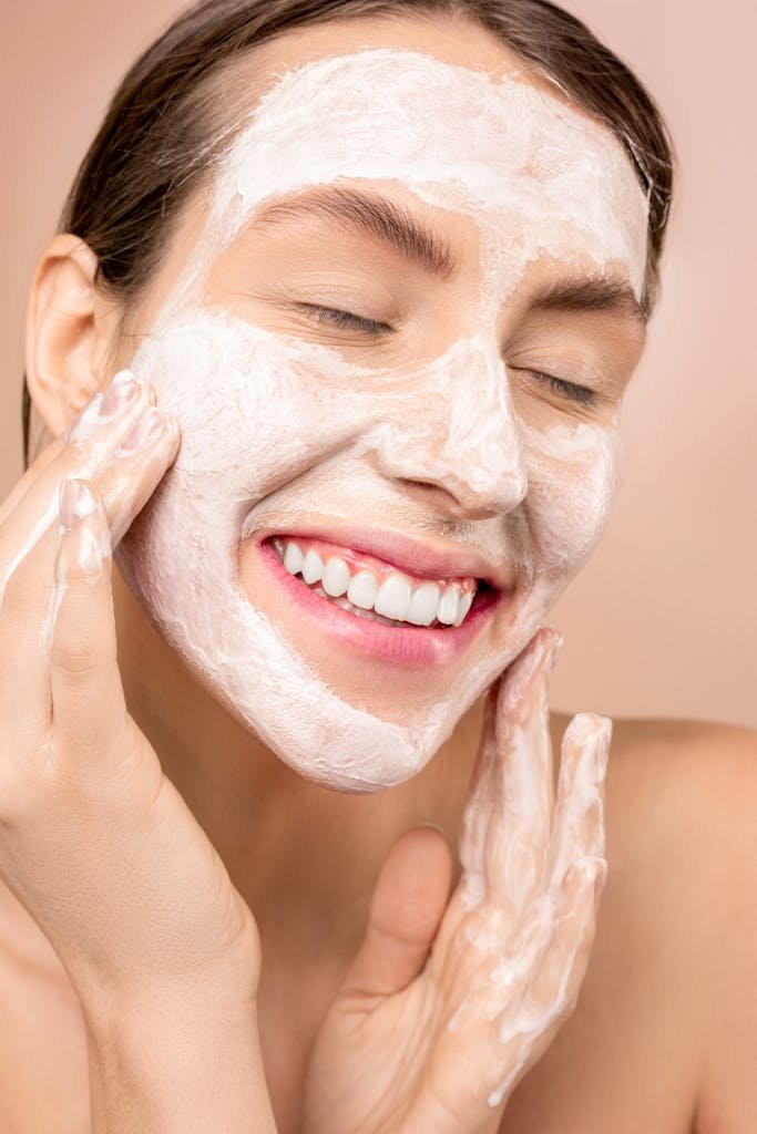 A woman with glowing skin smiling while applying facial cleanser for skincare routine.