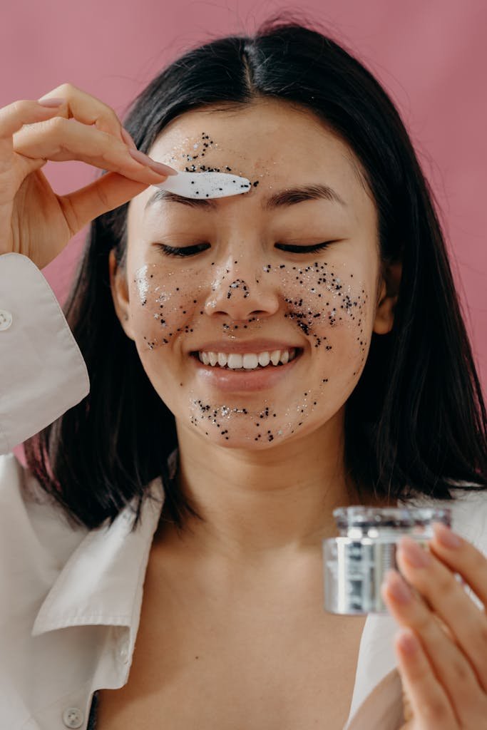 Smiling Asian woman applying an exfoliating face mask with a spatula.